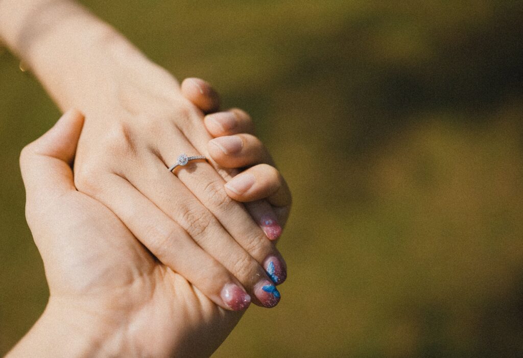 A close-up image of a hand wearing an engagement ring, with colorful nail design, in sunlight.